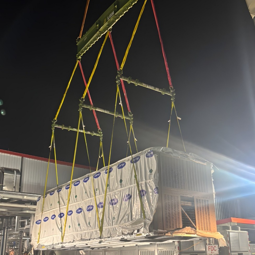 Crane lifting a wrapped Carrier HVAC unit onto a rooftop at night with engineered multi-leg slings