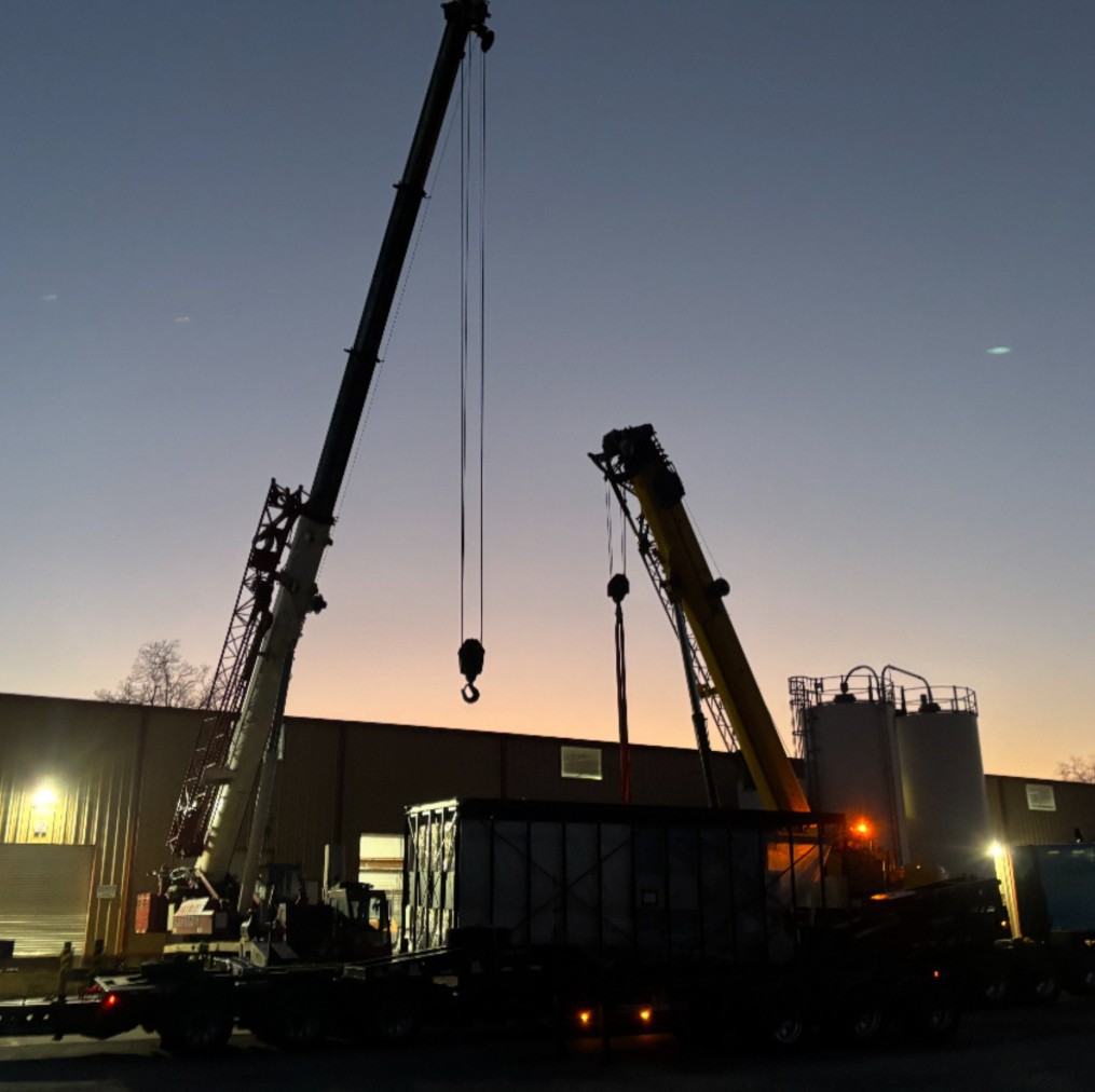 Two cranes silhouetted at dusk lifting an enclosed industrial structure