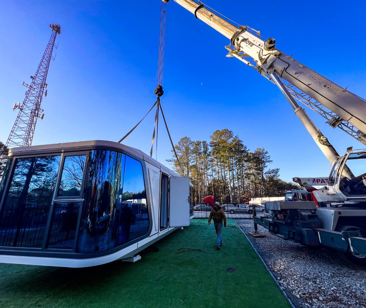 Crane lifting a modern modular Cocoon home with mirrored exterior next to a cell tower