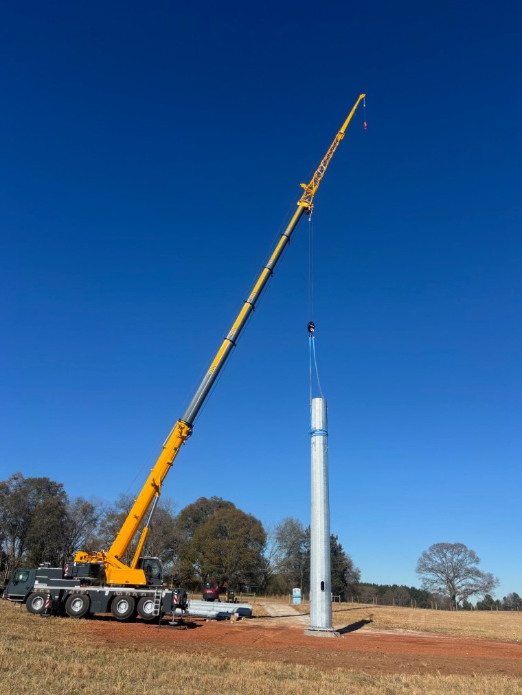 Yellow Liebherr all-terrain crane lifting a tall steel monopole tower in an open field