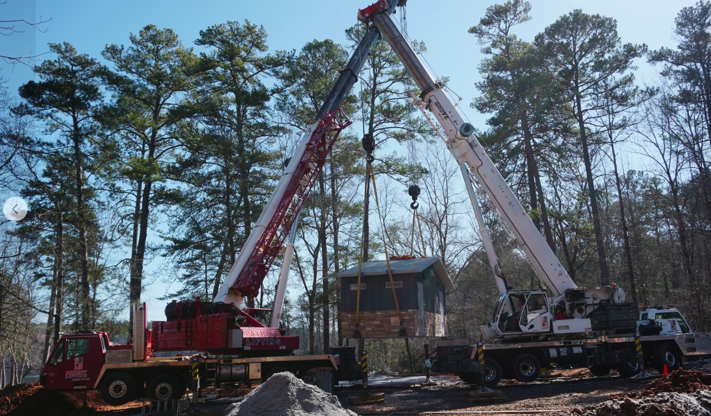 Two red cranes performing a tandem lift of a small modular structure in a wooded setting