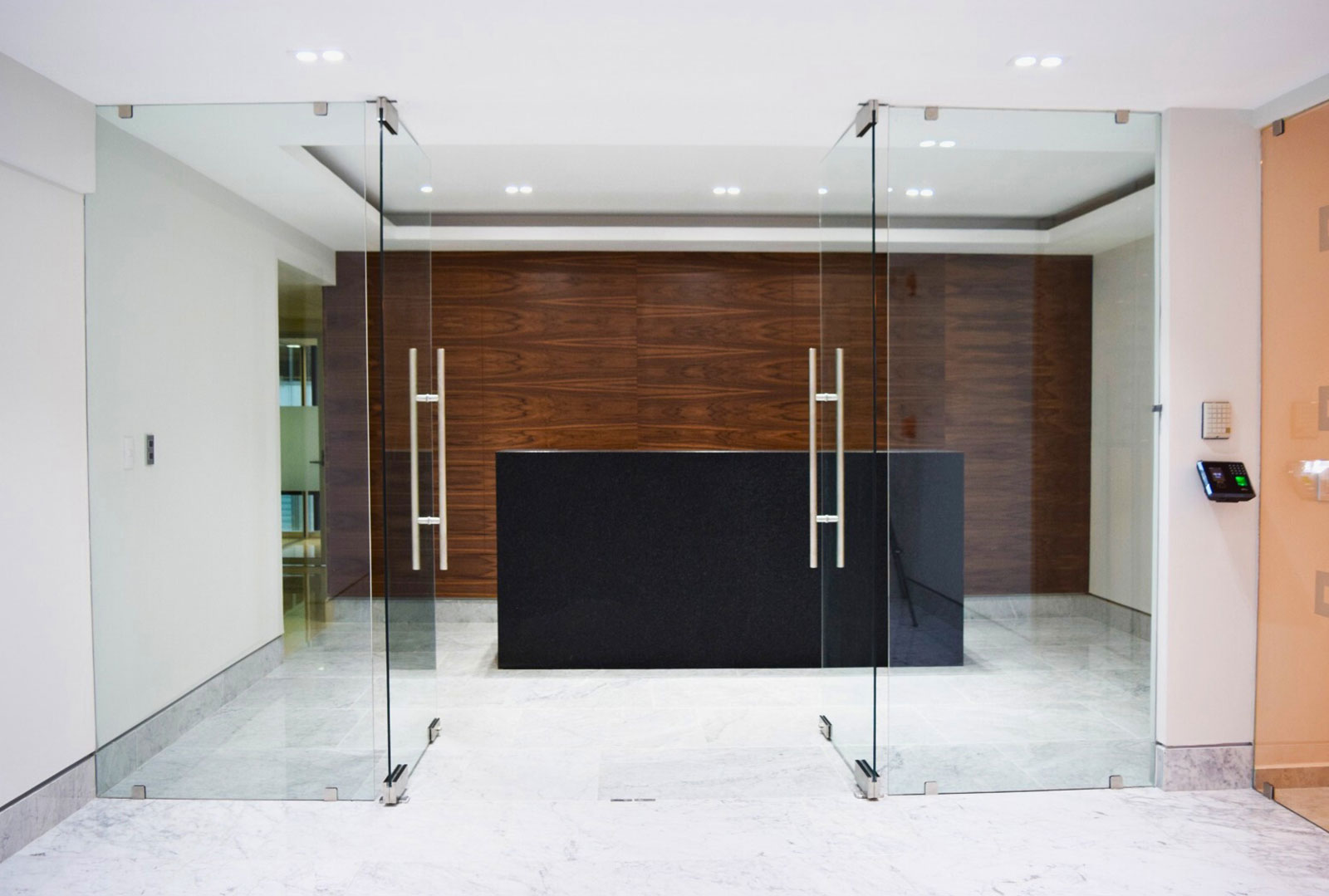 Modern office lobby with glass doors, black reception desk, wood panel wall, and marble floor.