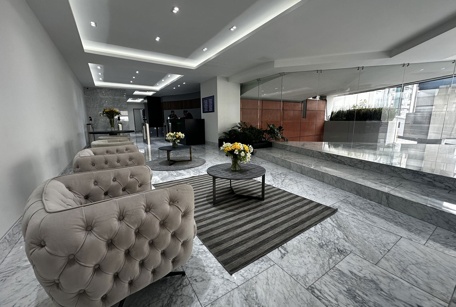 Modern lobby with tufted beige chairs, round tables with flower vases, and glass walls with city views.