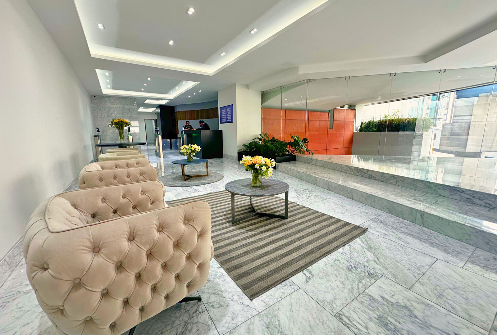 Modern lobby with beige tufted chairs, round tables with flower vases, marble floor, and glass wall.
