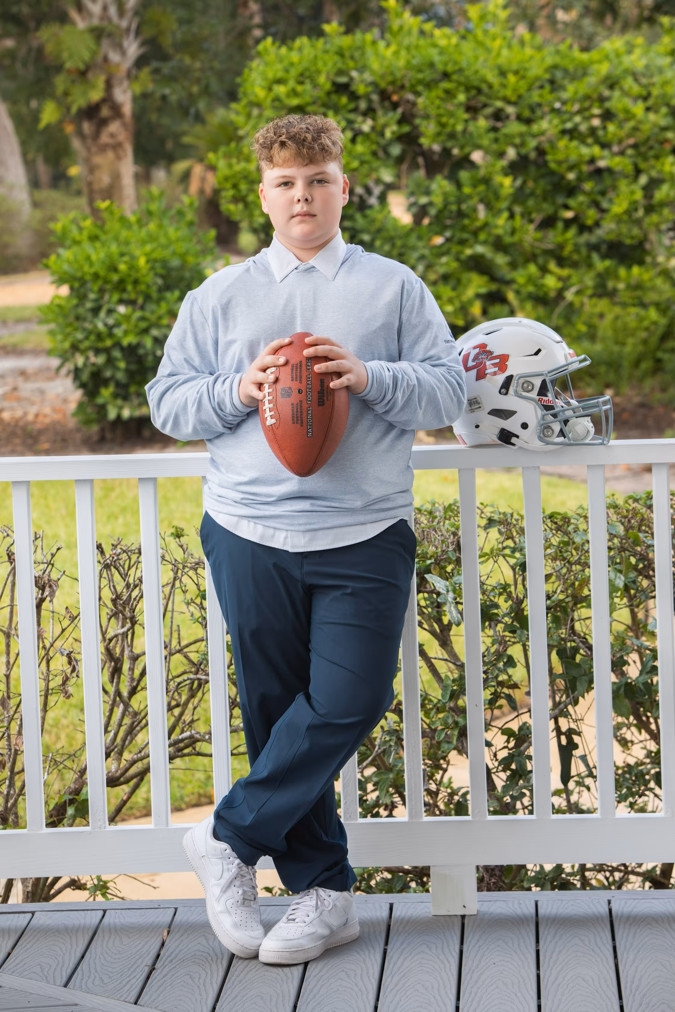 Young boy holding a football standing on a porch with a football helmet on the railing.