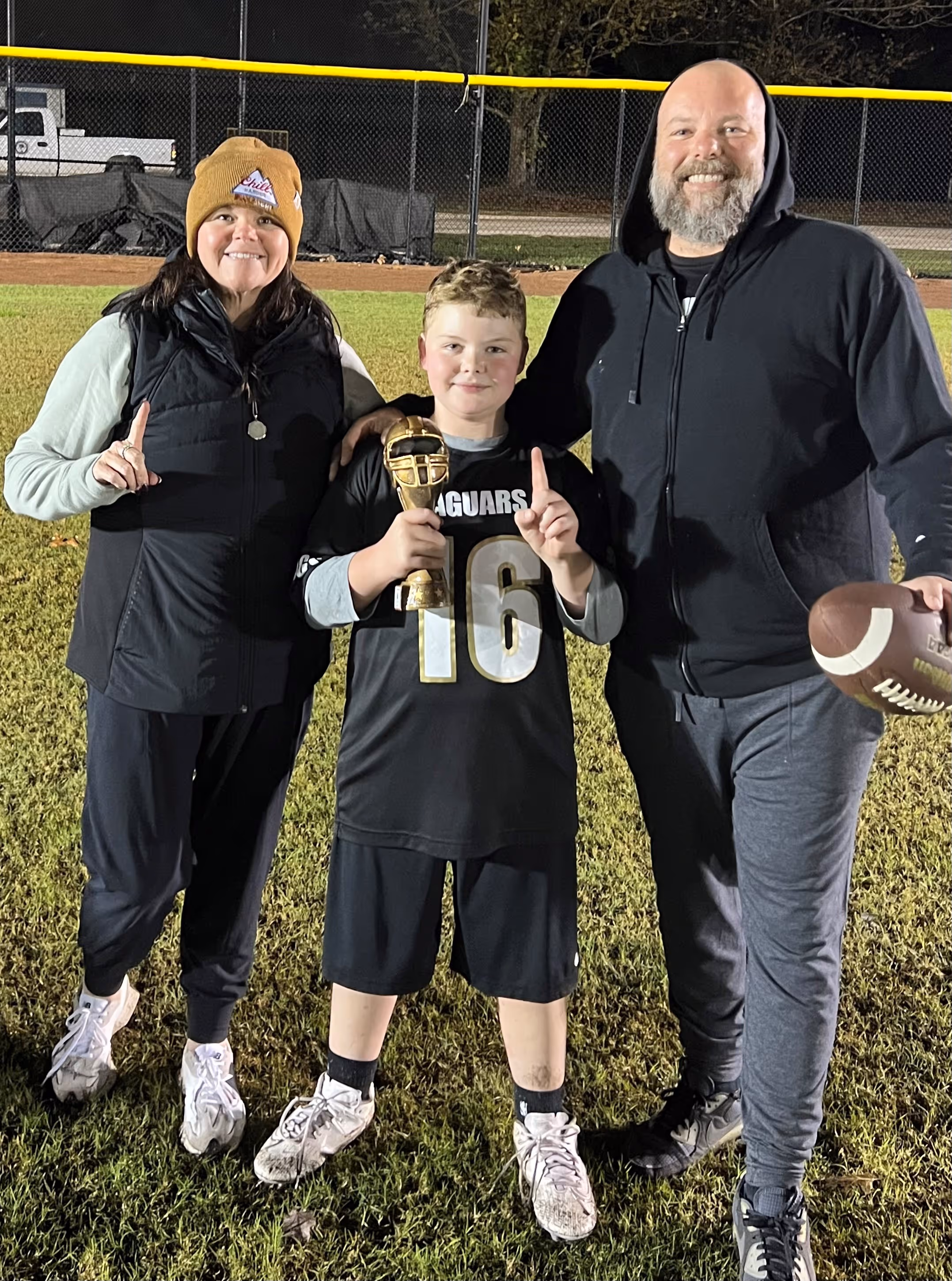 Smiling boy in football jersey holding trophy, flanked by two adults on a football field at night.