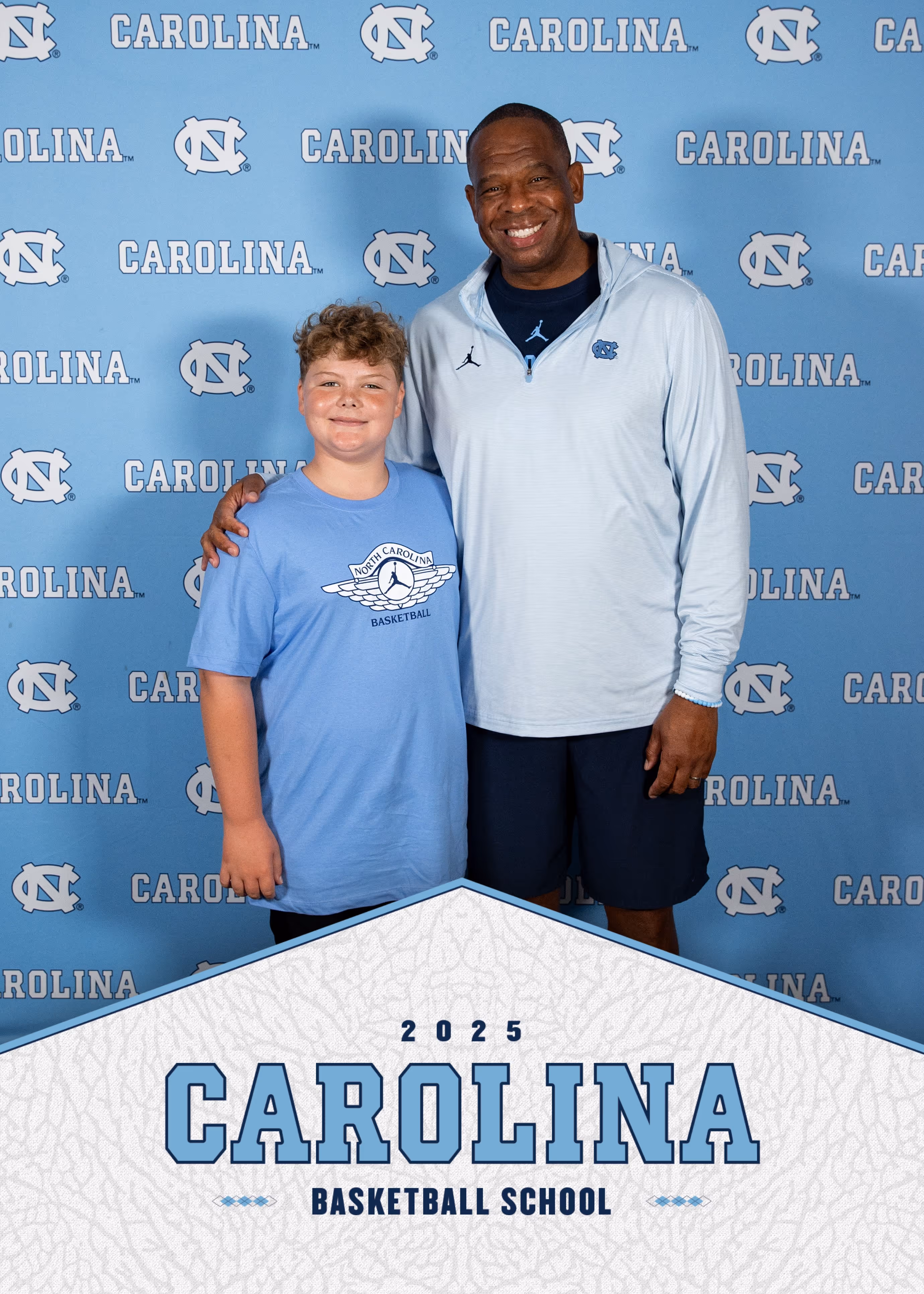 Smiling man and boy in blue shirts posing in front of a Carolina basketball backdrop.