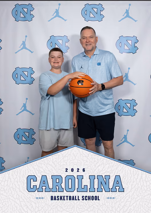 Two males in light blue shirts holding a basketball in front of Carolina Basketball School backdrop.