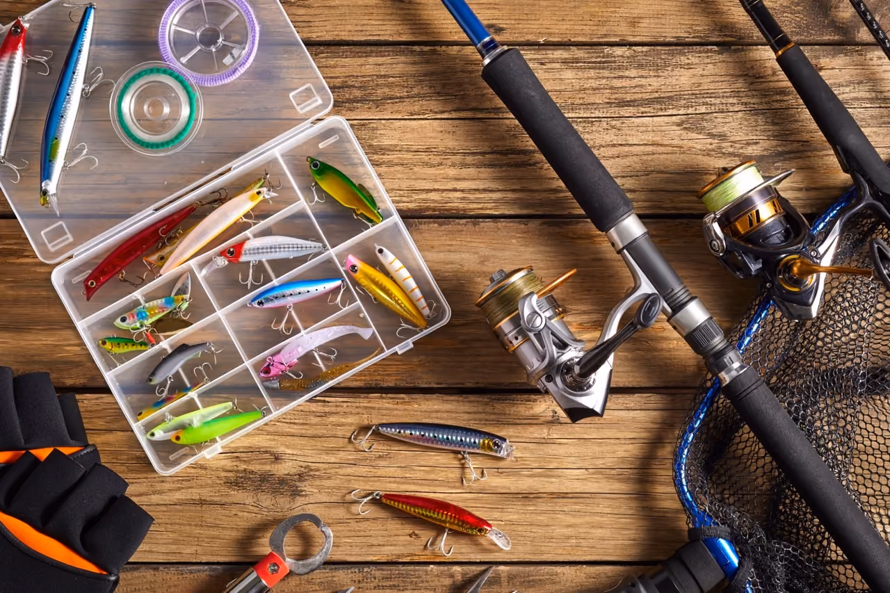 Fishing gear with rods, reels, colorful lures, gloves, and net on a wooden surface.