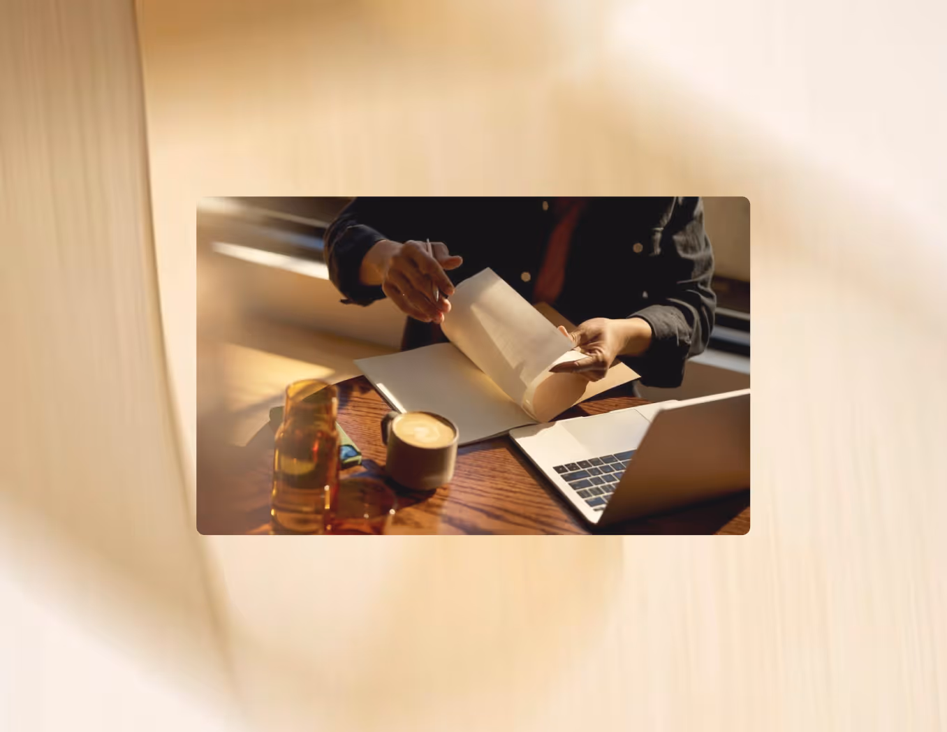Person flipping pages in a notebook with a laptop, coffee cup, and glass on a wooden table.