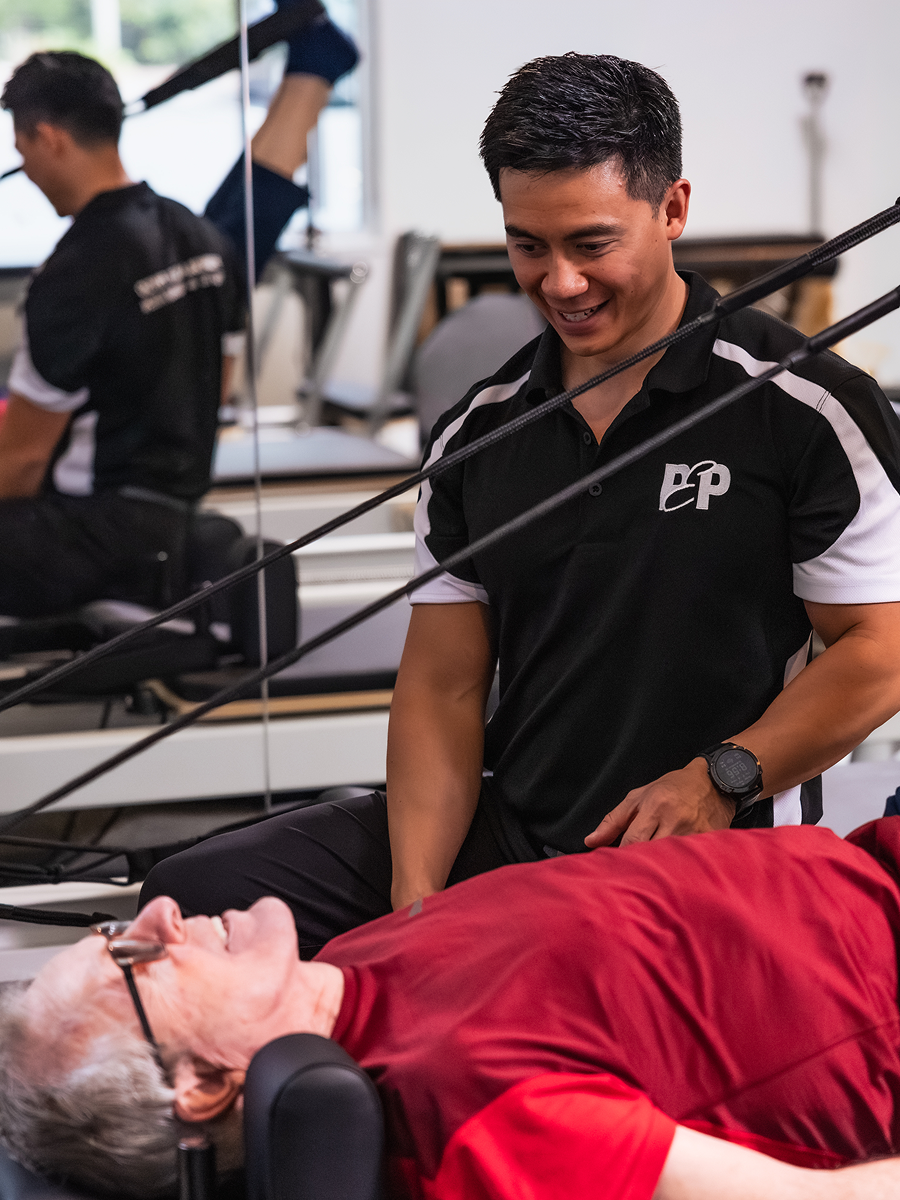 Male trainer assists elderly man doing rehabilitation exercises on a reformer machine.