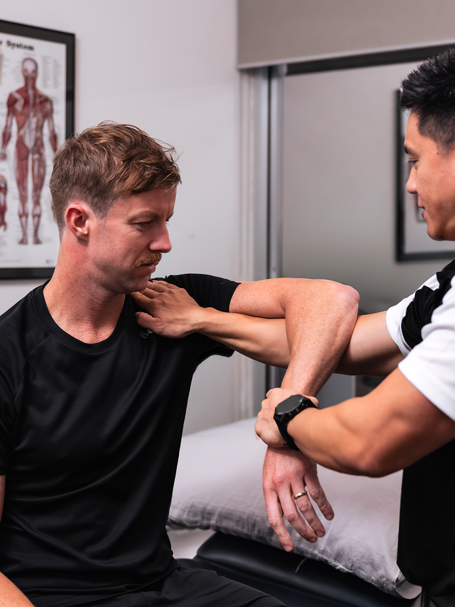 Physical therapist assists man with arm stretch during rehab in a clinic room with anatomy poster.