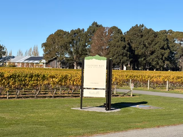 Sign reading 'Cloudy Bay New Zealand Closed Saké' in grass with vineyard and trees under clear blue sky.