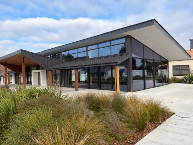 Modern building with large glass windows, black roof, wooden pillars, and landscaping in front.