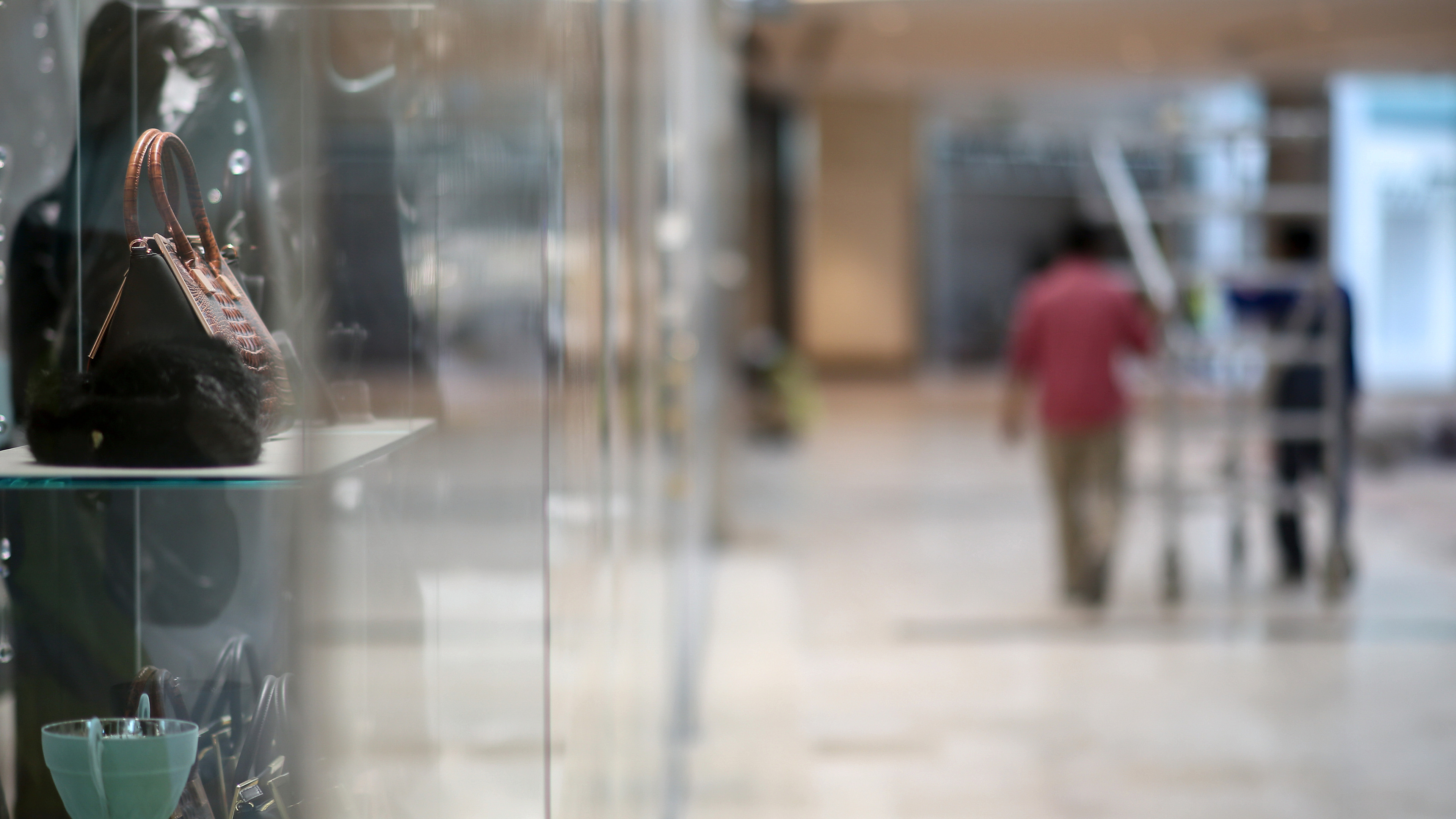 A handbag on a glass shelf in a shopfront window, with a shopper passing on the concourse beyond.