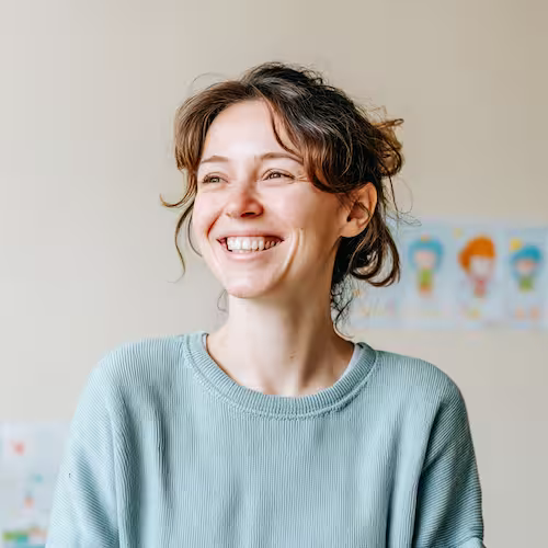 Smiling woman with brown hair in a loose bun wearing a light blue sweater looking to the side indoors.