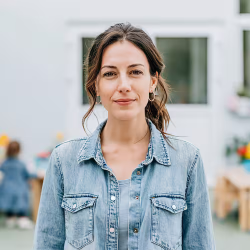 Woman with brown hair in a ponytail wearing a denim jacket and hoop earrings, smiling slightly outdoors.