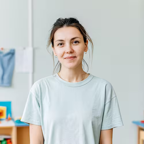 Smiling woman with tied-back hair wearing a light blue t-shirt in a softly blurred indoor setting.