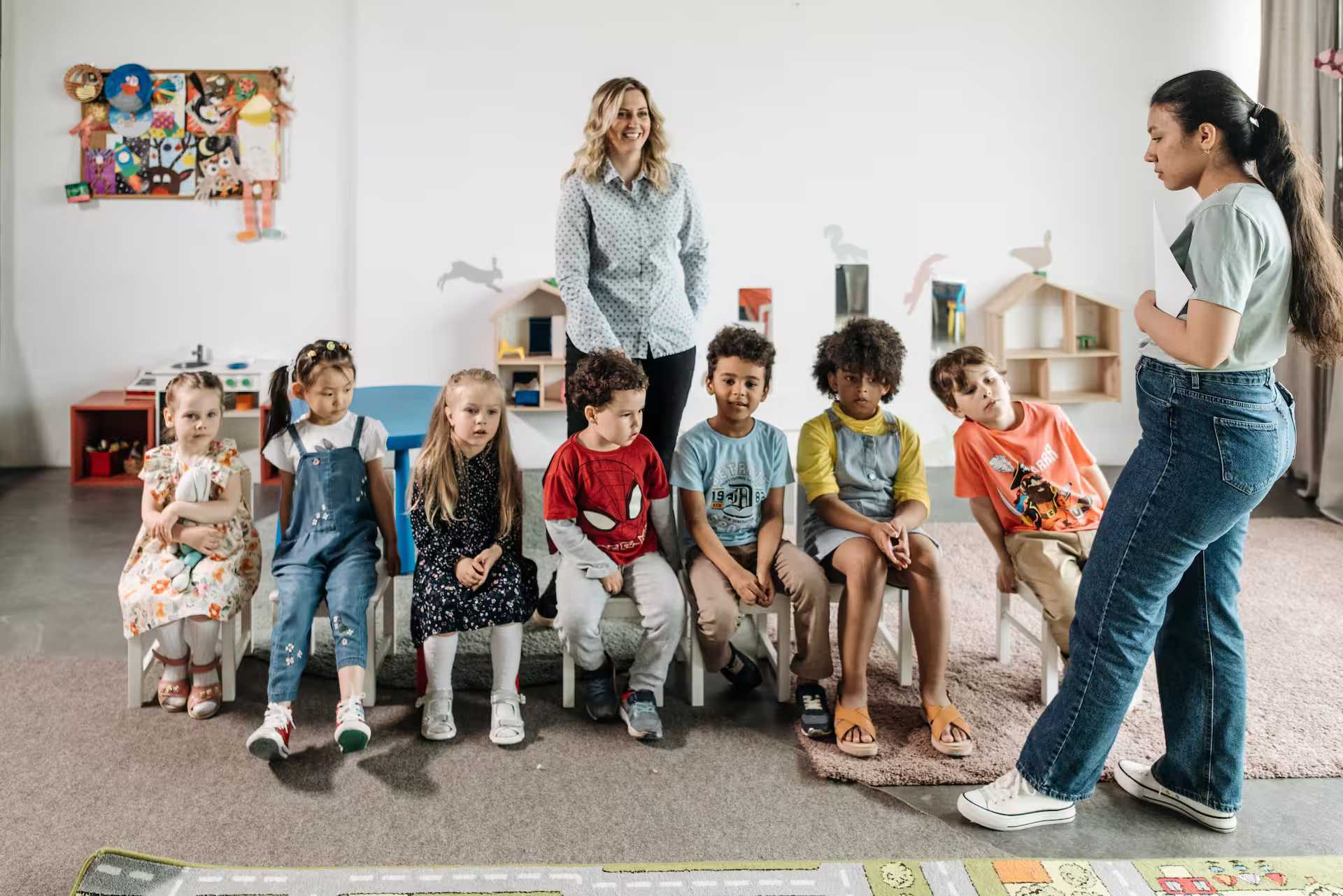 Seven children sitting on chairs in a classroom with two female teachers standing nearby.