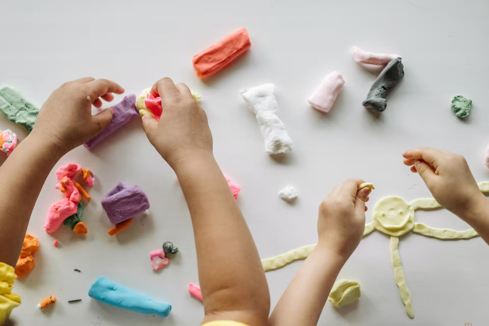 Child's hands shaping colorful modeling clay on a white surface, including a sun figure with a smiley face.