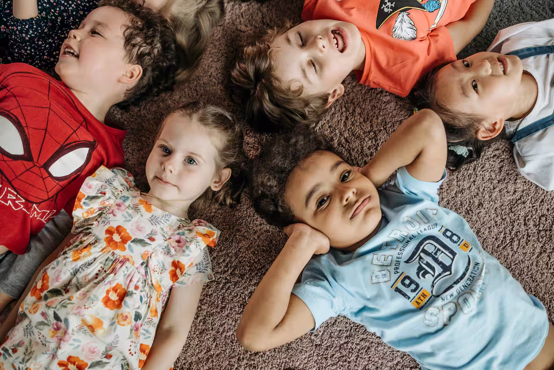Six diverse children lying in a circle on a carpet, smiling and relaxed.