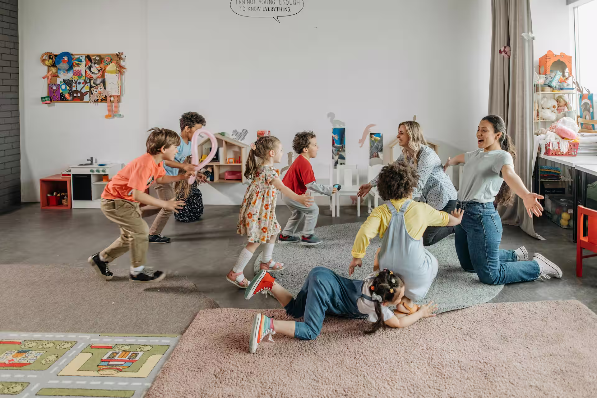 Children playing and laughing indoors with two adults in a bright, colorful playroom.