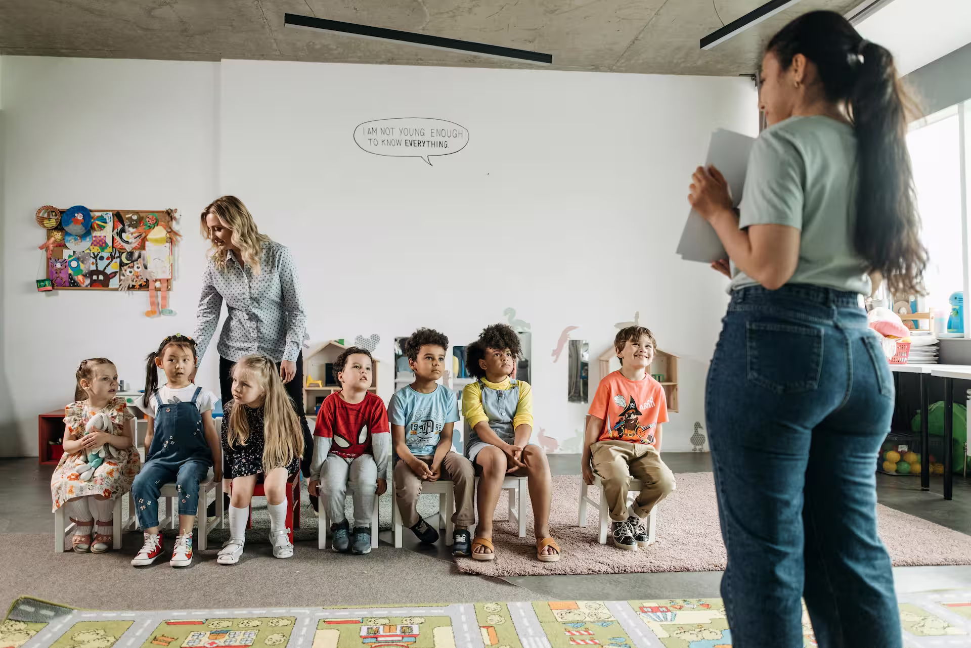 Teacher standing in front of a seated diverse group of young children in a classroom.