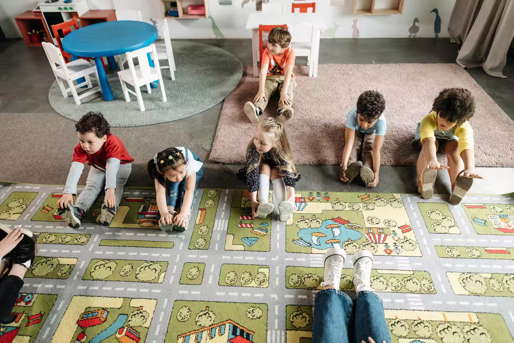 Group of children sitting on carpet stretching forward, following adult's lead in a classroom.