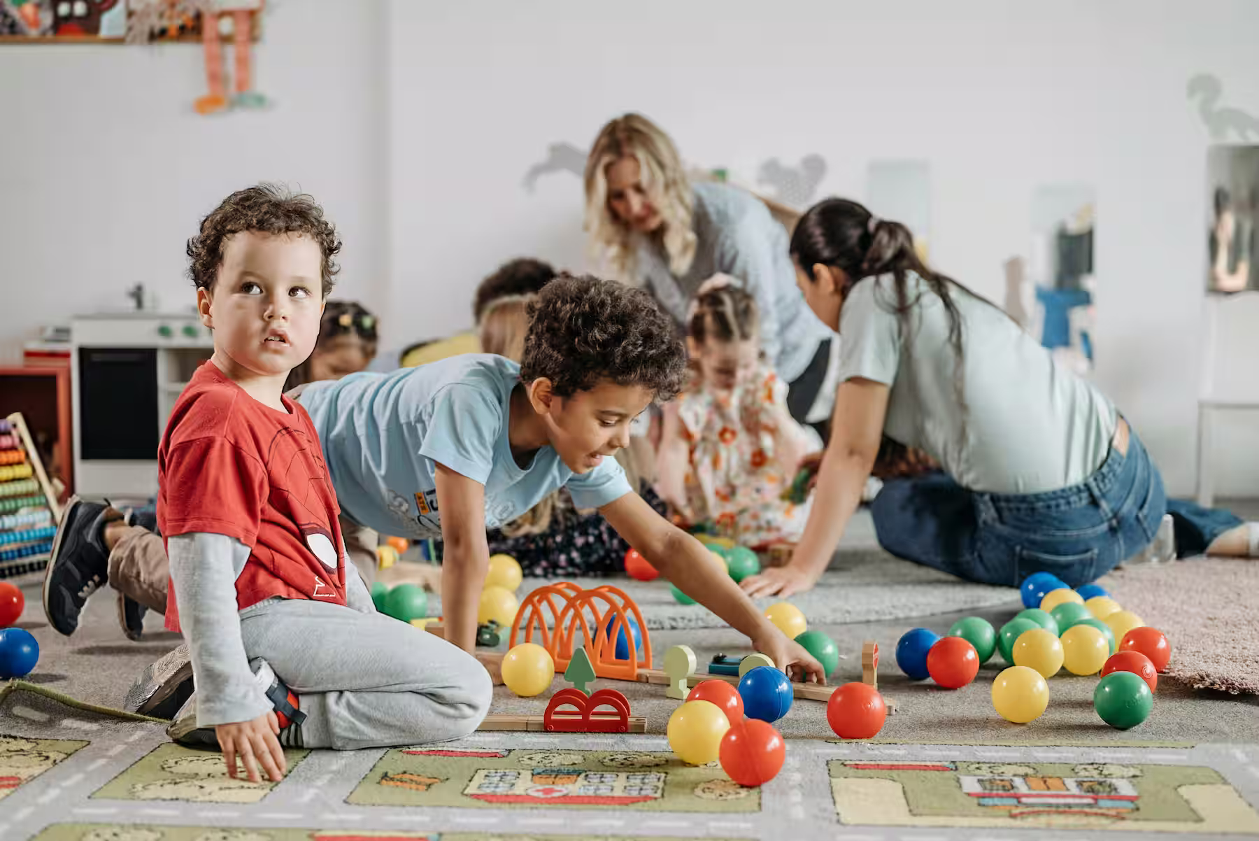Children playing with colorful balls and toys on a carpet in a bright playroom with two adults.
