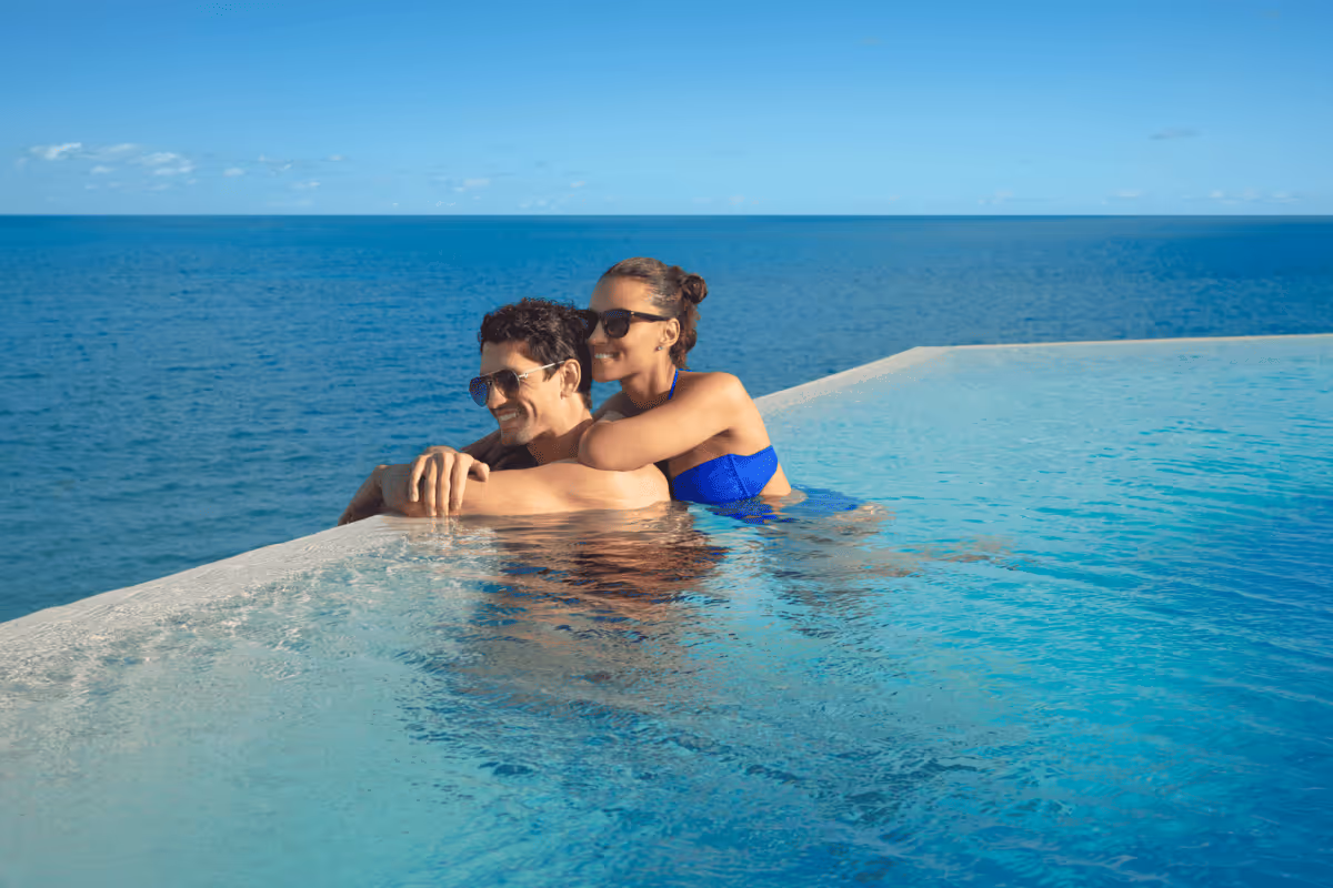 Couple wearing sunglasses relaxing in an infinity pool overlooking the ocean on a sunny day.