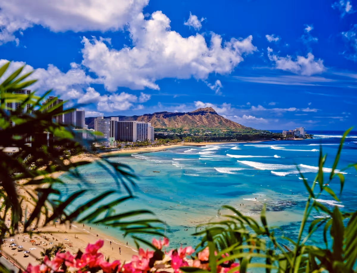 View of a tropical beach with turquoise water, city buildings, mountains, and a partly cloudy blue sky.