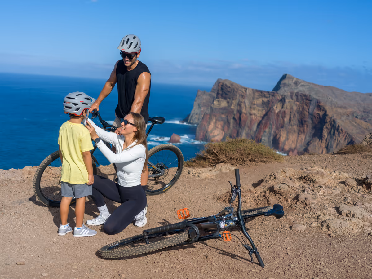 Woman helps child with bike helmet while man with bike watches on cliff by ocean and rocky mountains.