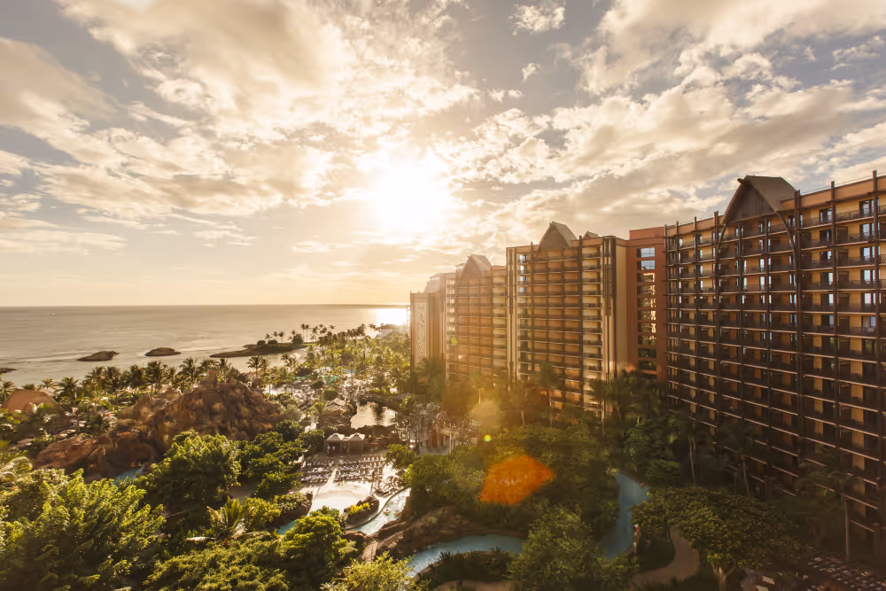 Sunset over a tropical resort with tall buildings, palm trees, pools, and ocean in the background.