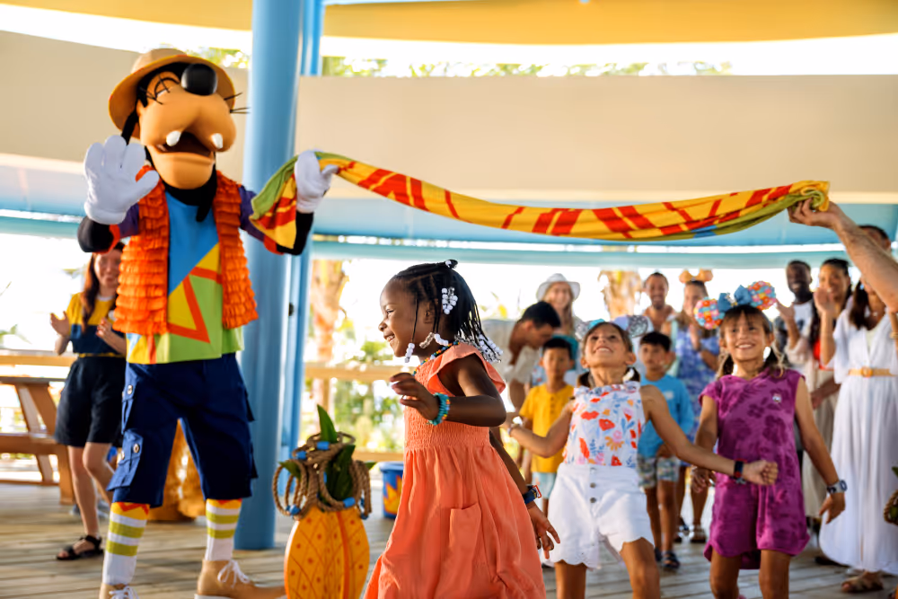 Children play limbo under a colorful scarf held by a person in a Goofy costume at a festive event.