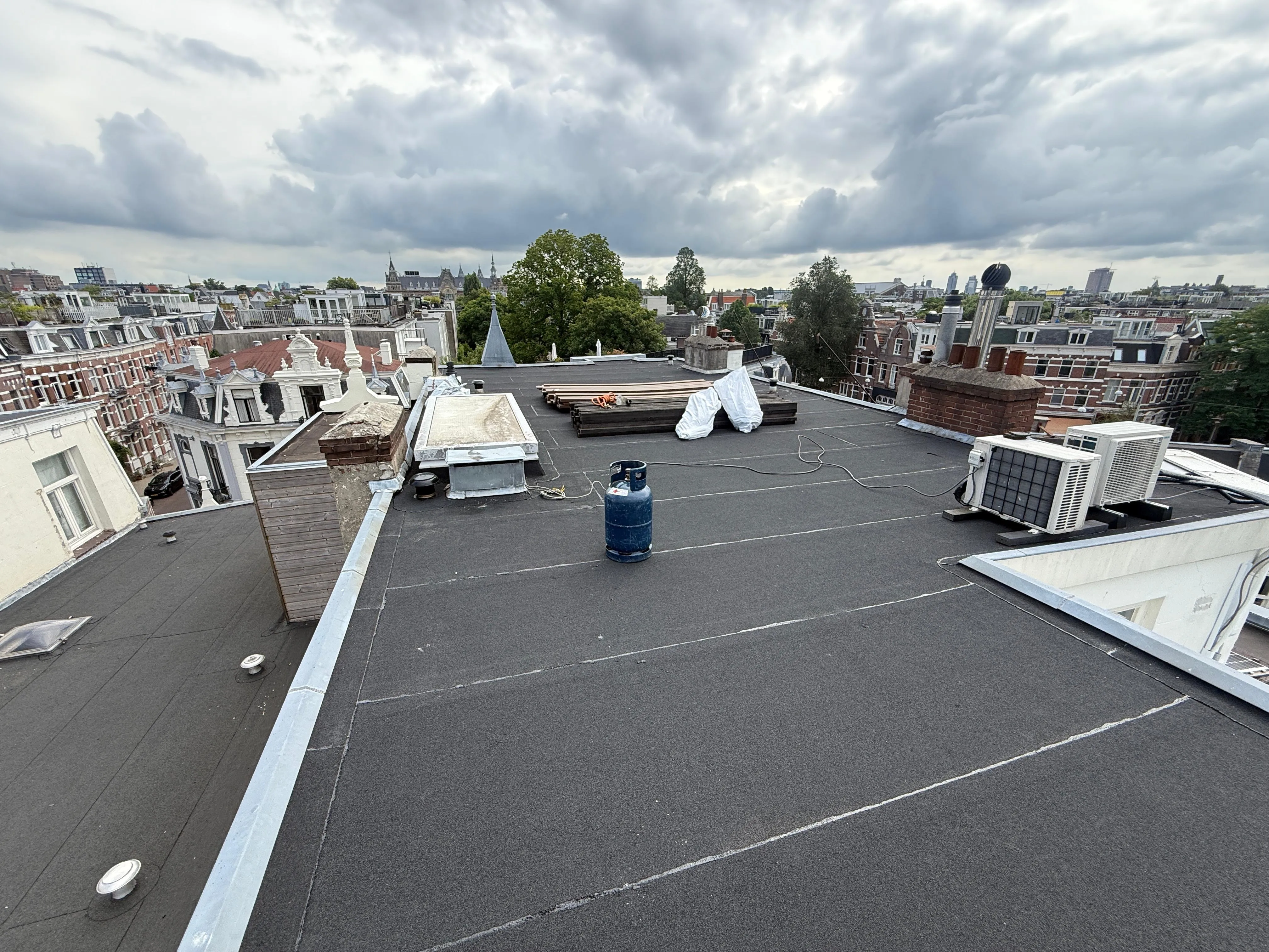 Plat dak in Amsterdam met airconditioners, gasfles en bouwmaterialen onder bewolkte lucht.