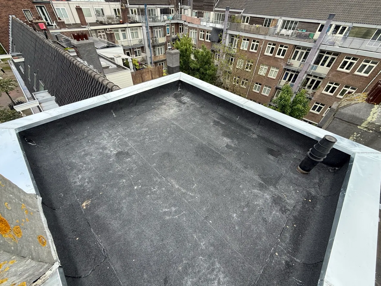 Plat dak met scheuren en lichte mosvorming, omringd door stedelijke woongebouwen.