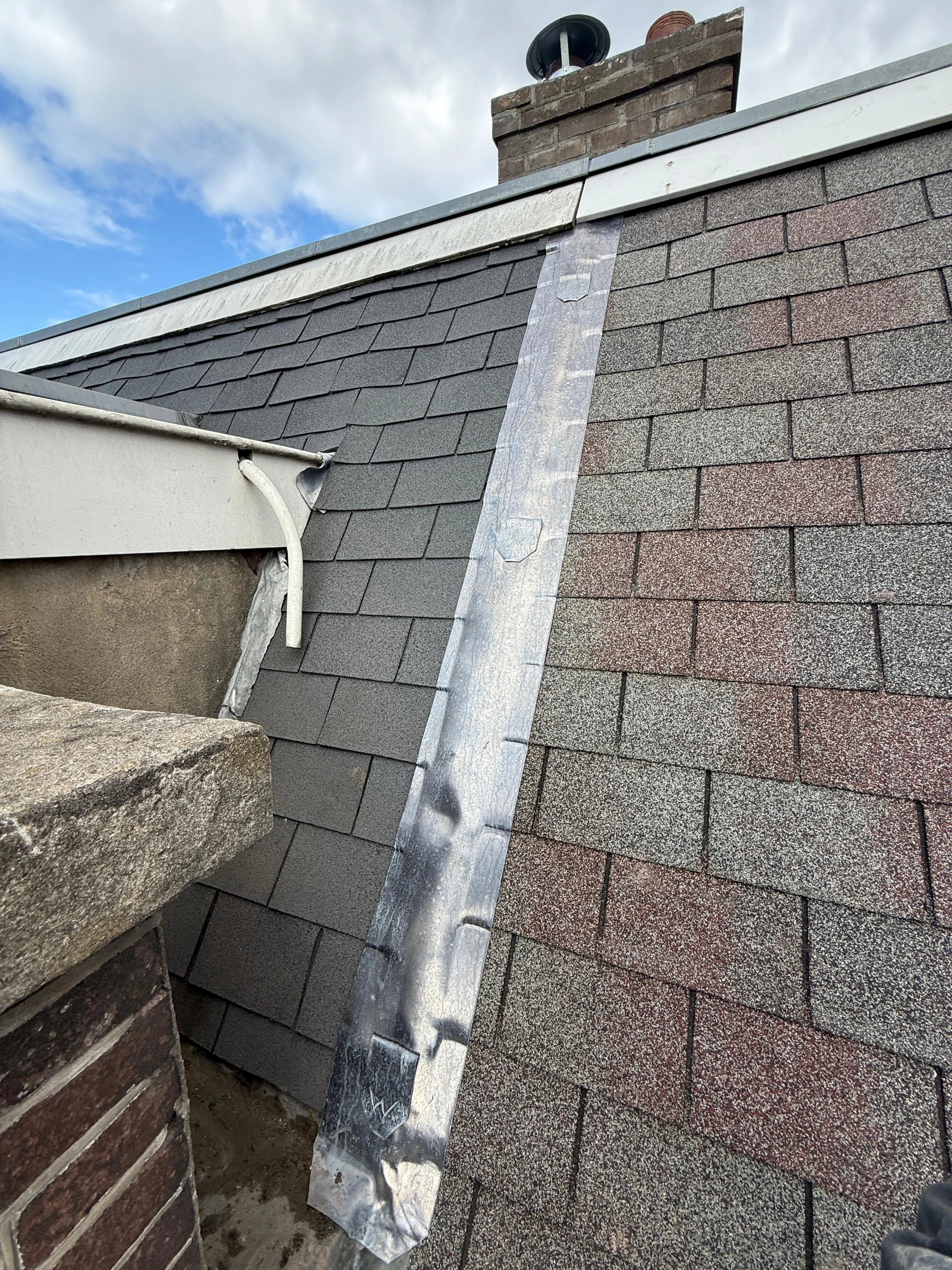 Dak met nieuwe en oude dakshingles naast een metalen dakgoot langs een schoorsteen.