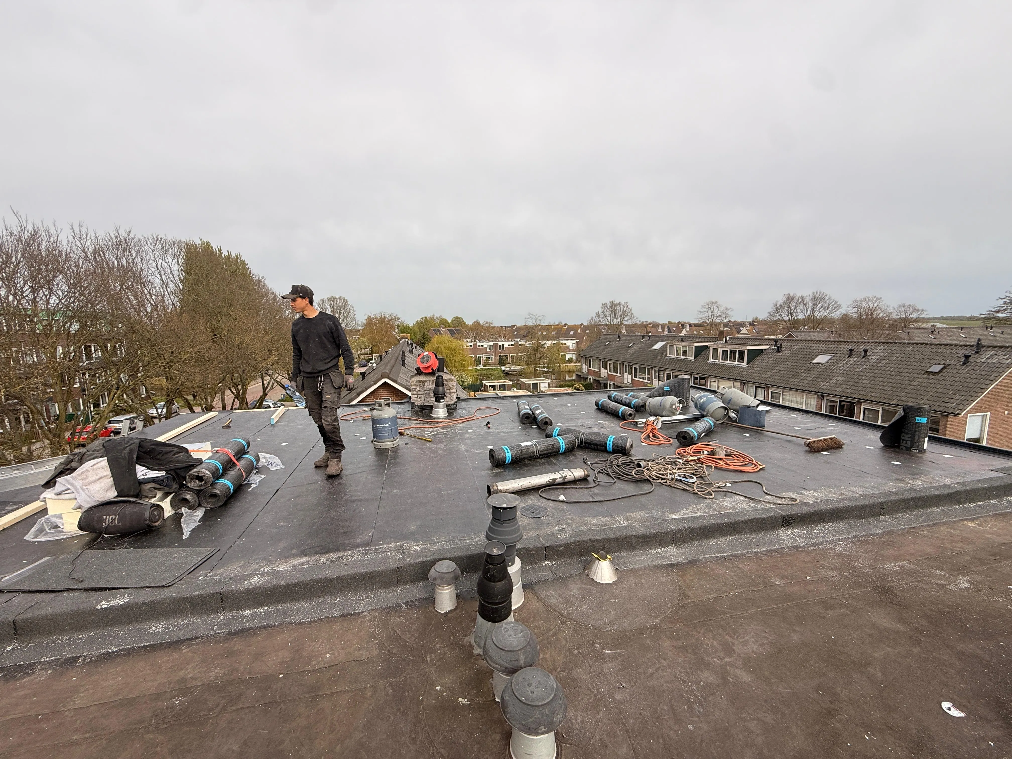 Dakrenovatie met arbeider, rollen dakmateriaal en gereedschap op plat dak in woonwijk.