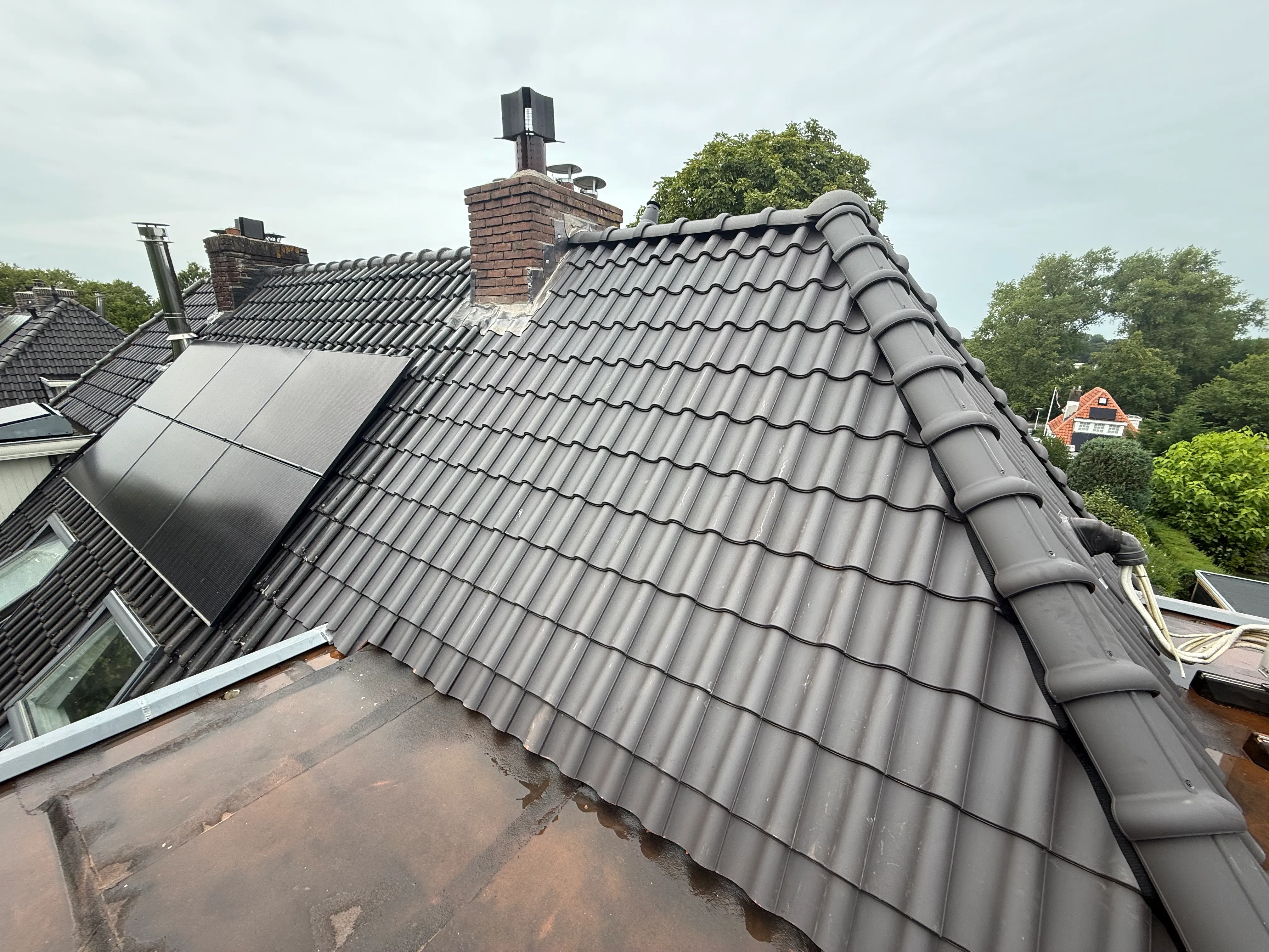 Dak met zwarte zonnepanelen naast grote schoorsteen en grijze dakpannen onder bewolkte lucht.