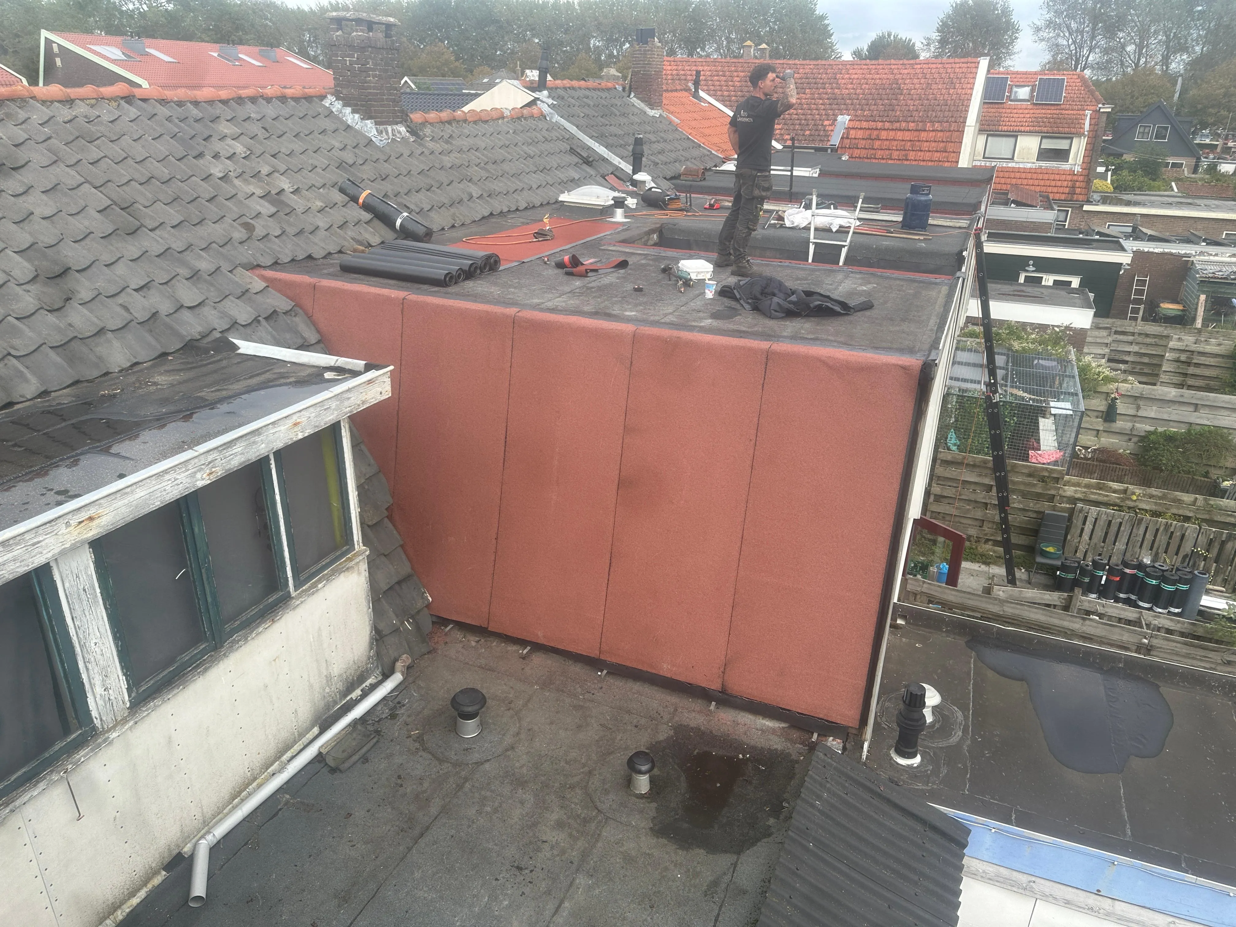 Dakwerker die een plat dak met rode membraan aan het herstellen is op een huis met omliggende daken.
