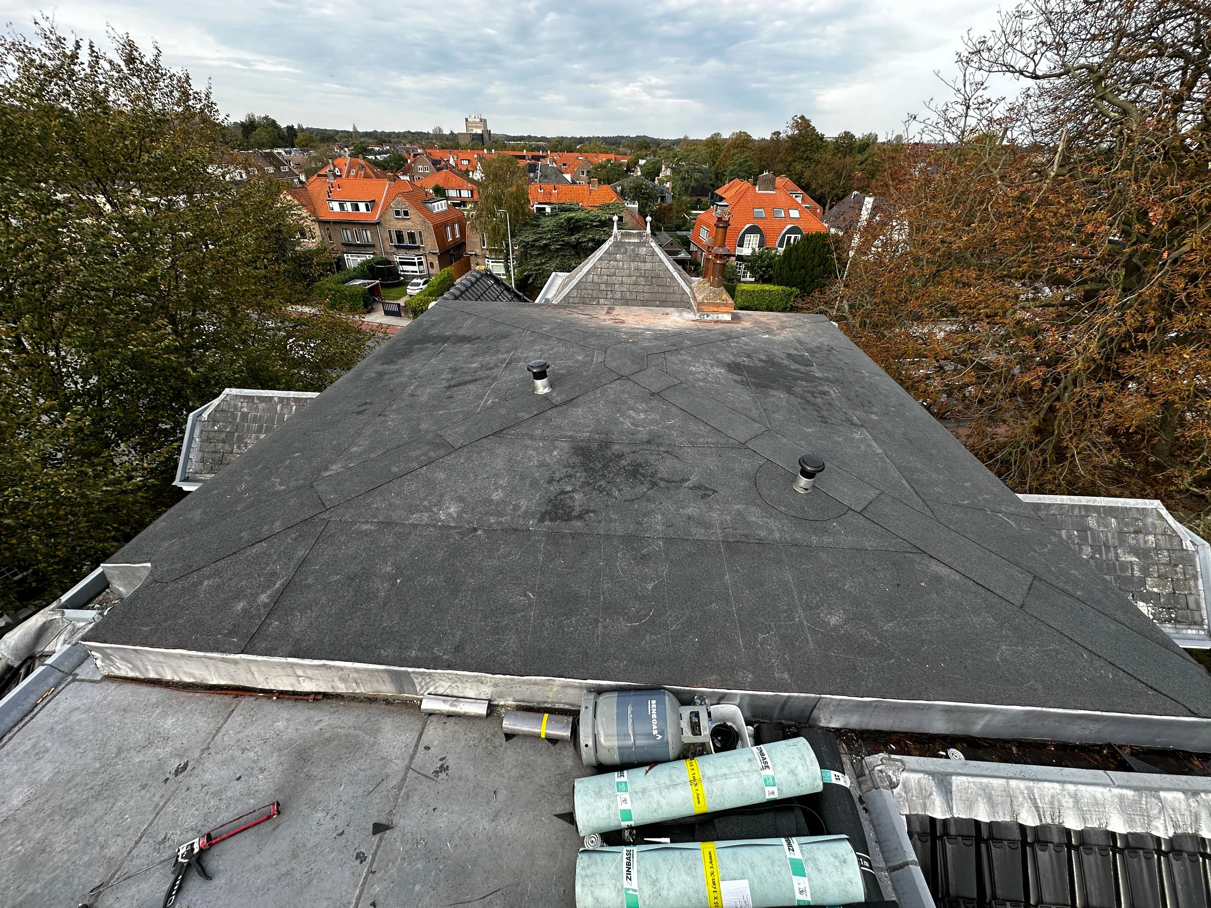 Dak met zwarte dakbedekking, gereedschap en rollen materiaal, omringd door huizen met rode daken.