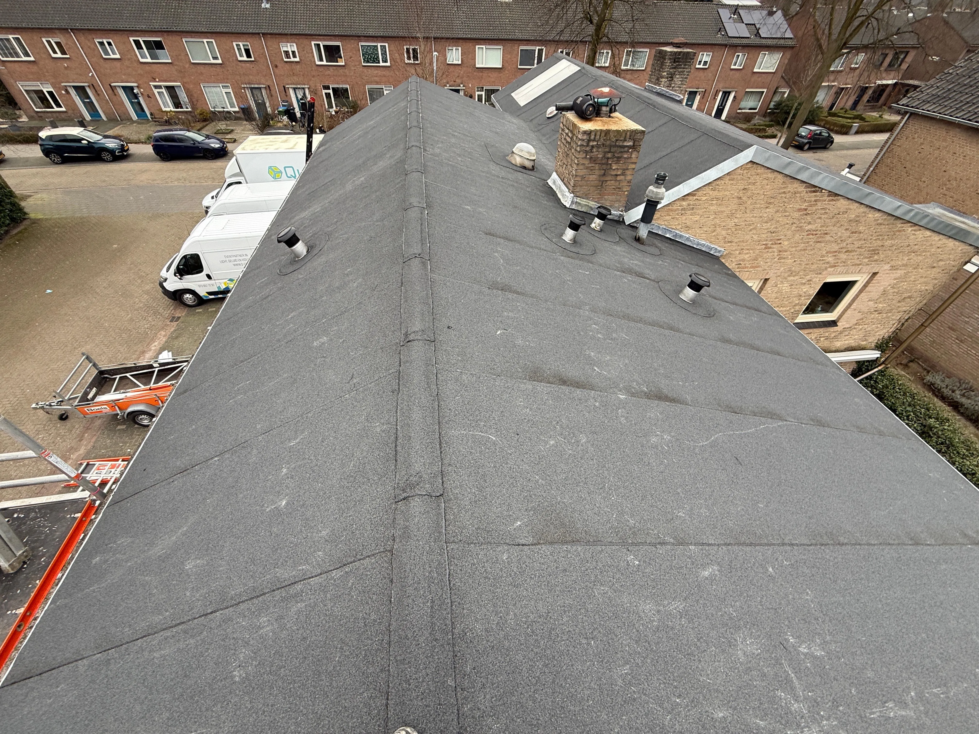 Grijs plat dak met schoorsteen en ventilatiepijpen gezien vanaf een verhoogd standpunt.
