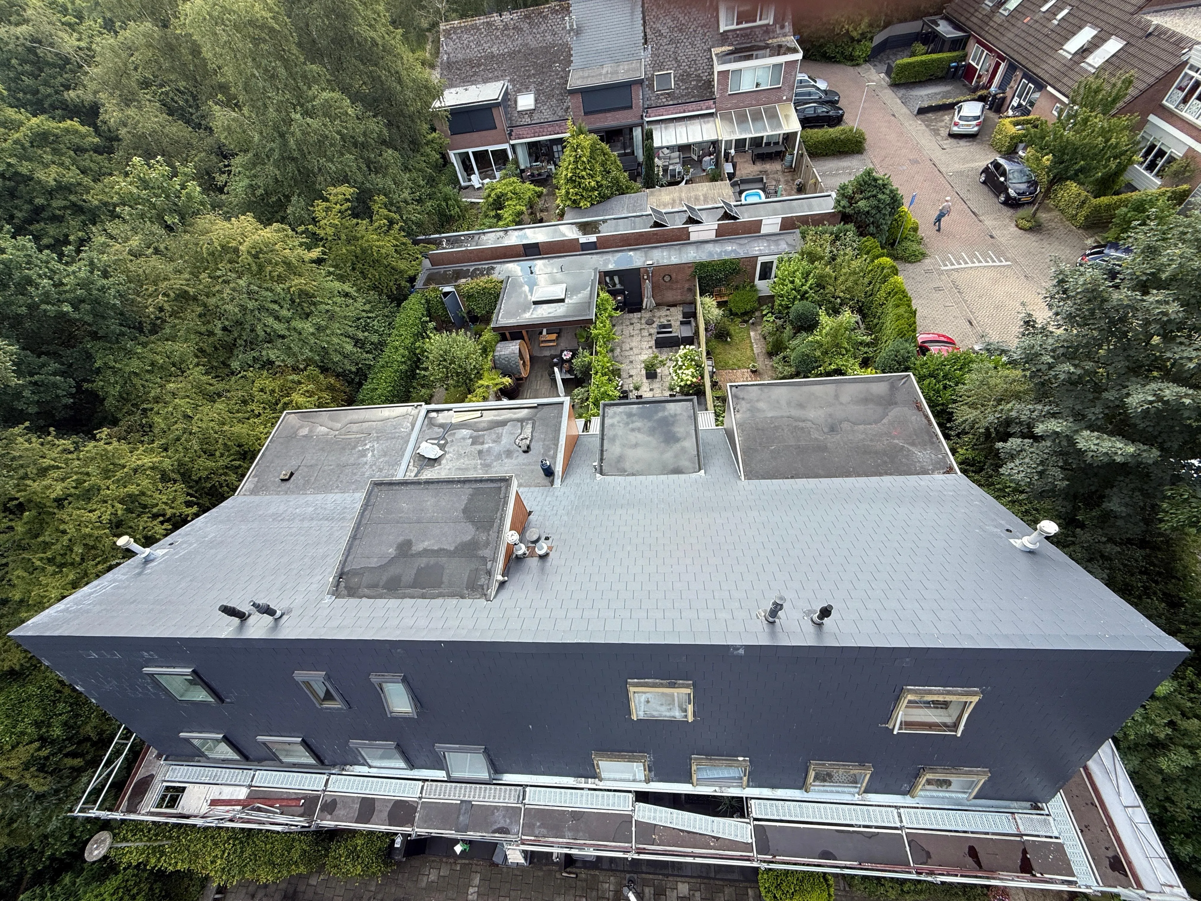 Luchtfoto van een gebouw met daken en tuinen, omgeven door bomen en een woonwijk.