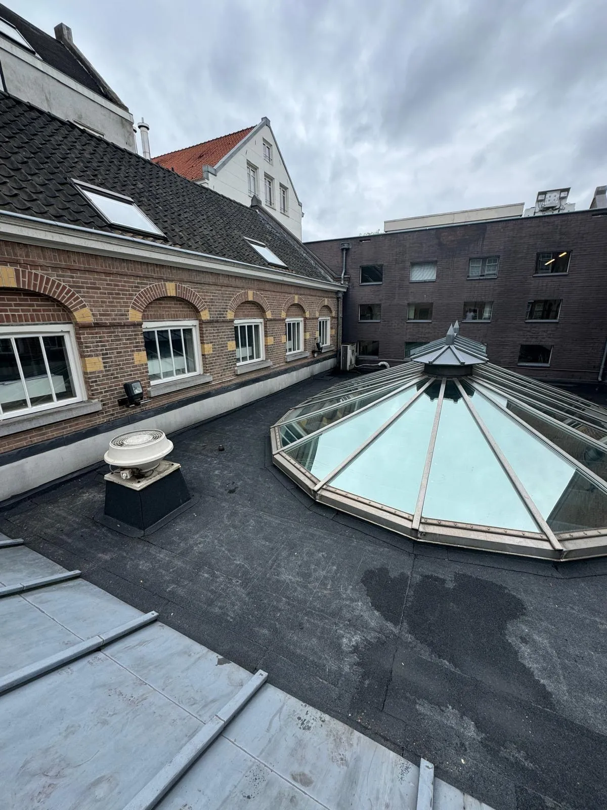 Dak met glazen koepel, ventilator en bakstenen gebouwen onder een bewolkte lucht.