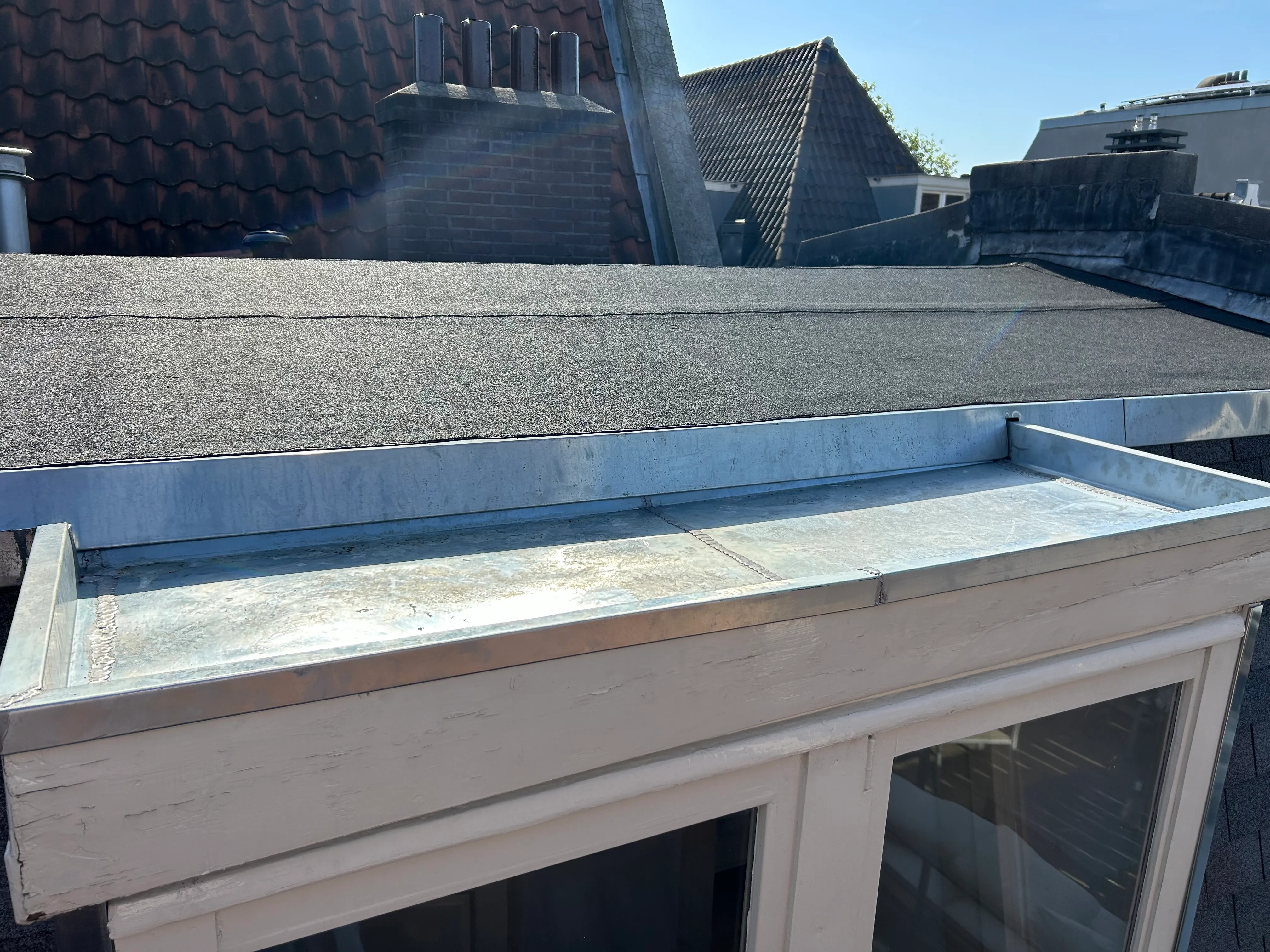 Dak met metalen dakrand en ramenolicht, omliggende dakpannen en schoorsteen zichtbaar.