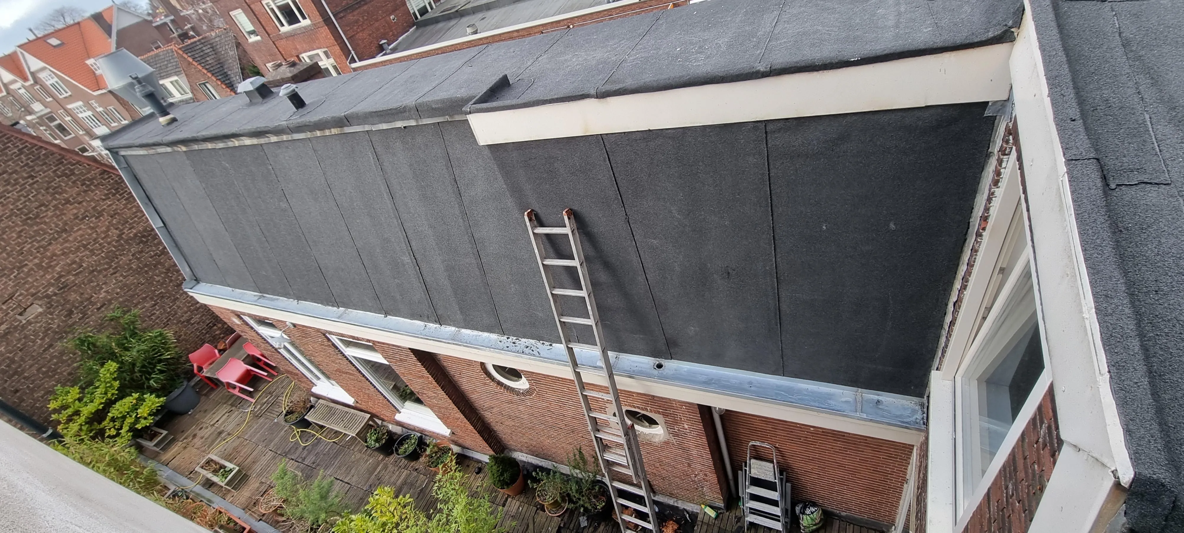Bakstenen gebouw met plat dak, metalen ladders en een beneden terras met planten en stoelen.