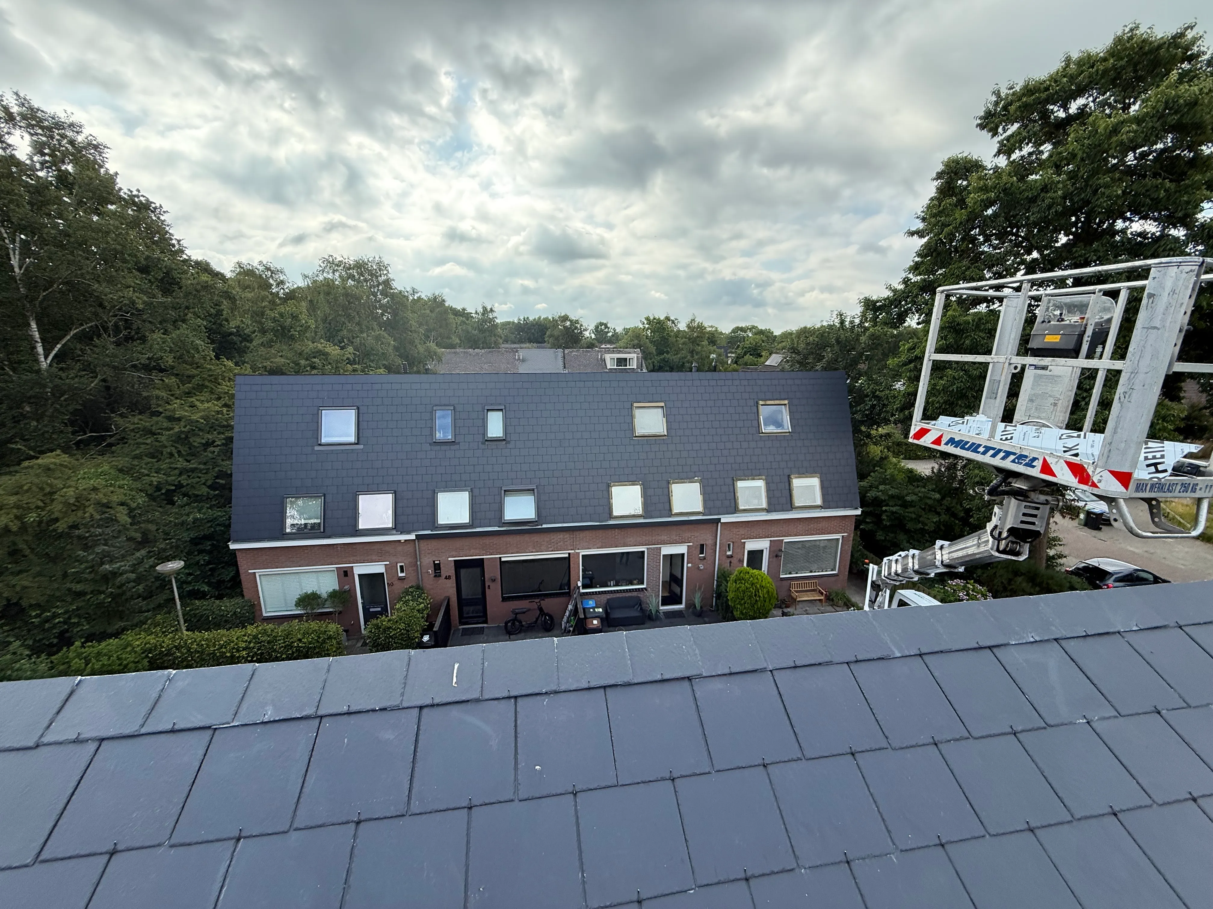 Rijtjeshuis met grijs dak, meerdere dakramen, groene bomen en een liftplatform aan de rechterkant.