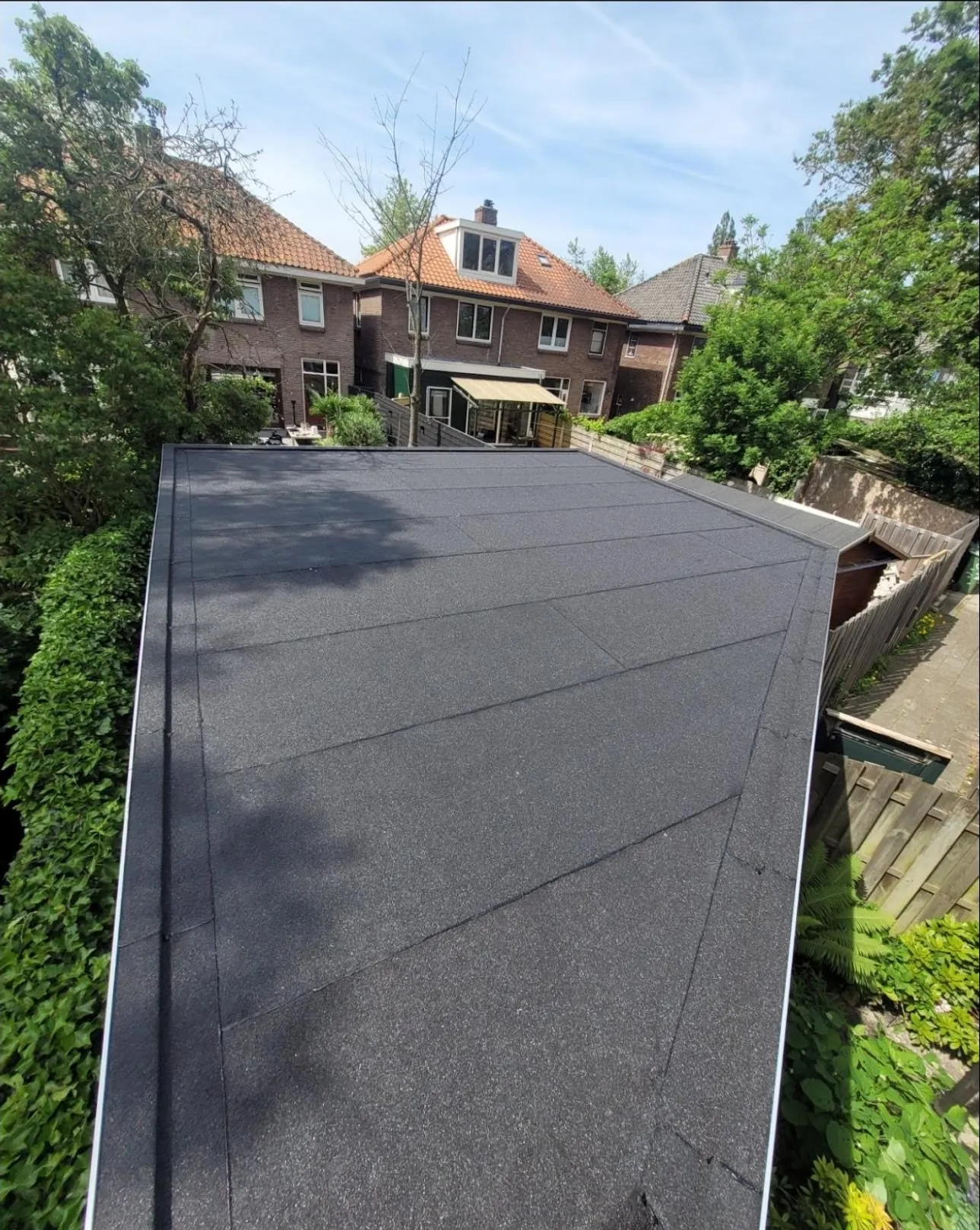 Plat dak bedekt met zwarte dakbedekking, omringd door groene struiken en huizen.