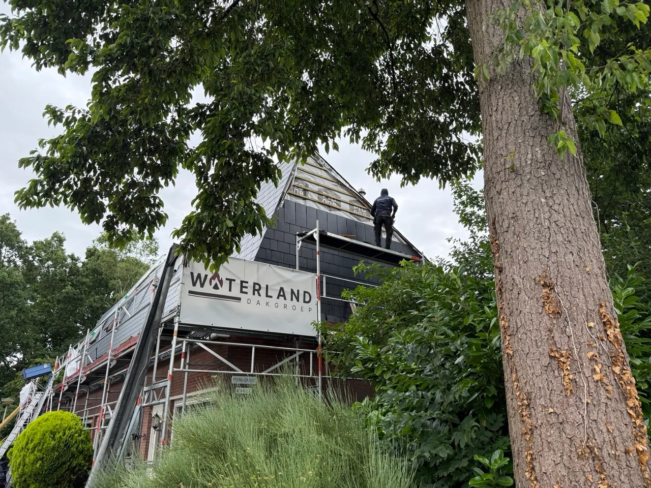 Bouwvakker op steiger plaatst dakpannen op een huis omgeven door bomen en struiken.