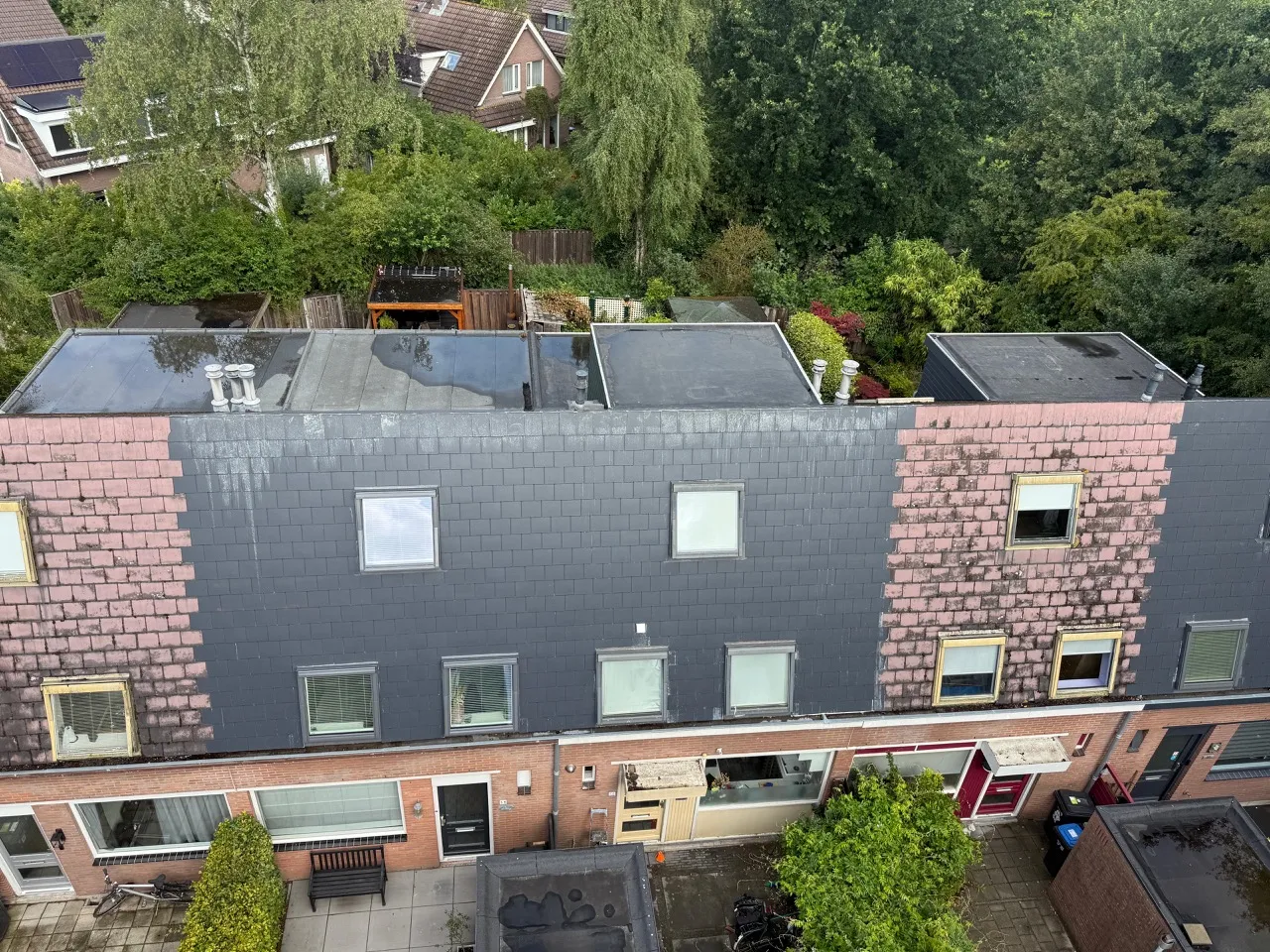 Rijtjeshuis met deels nieuwe grijze en deels oude roze dakpannen, omringd door bomen en tuinen.
